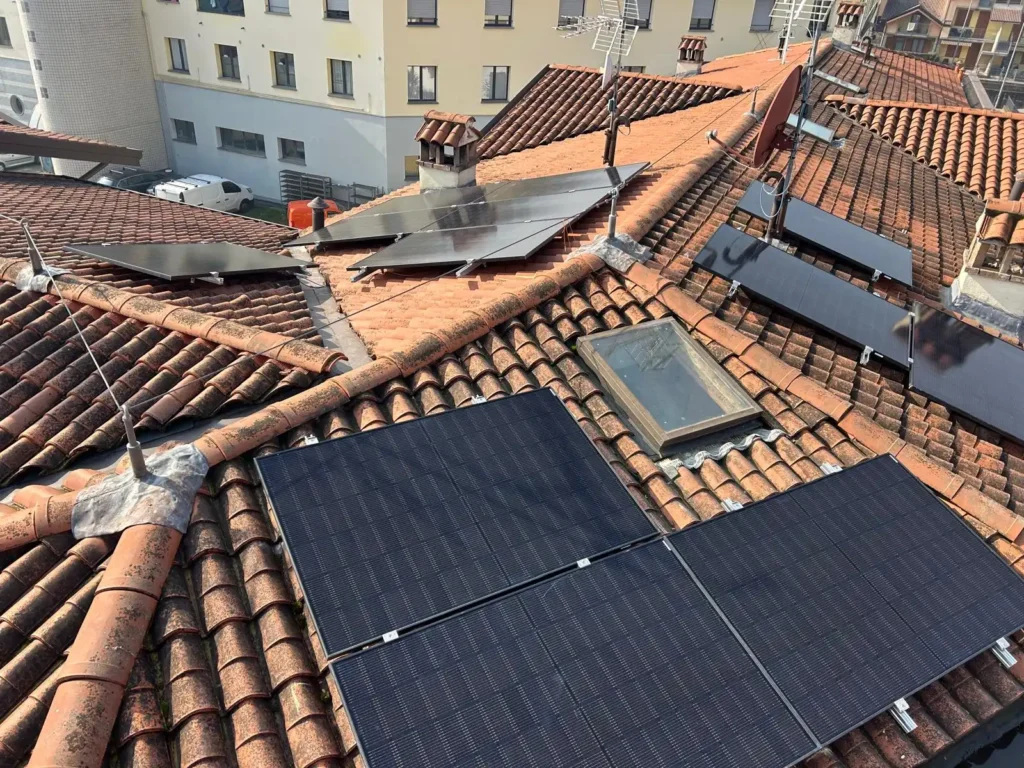 Un tetto a tegole rosse con diversi gruppi di pannelli solari neri e un lucernario, oltre a un'antenna televisiva e un paesaggio urbano sullo sfondo.
