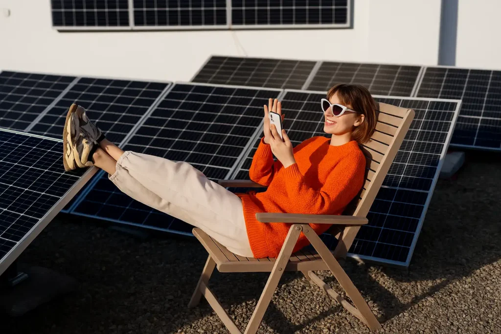 Una donna sorridente con un maglione arancione e occhiali da sole bianchi è seduta su una sedia a sdraio tra pannelli solari, mentre guarda il suo smartphone.