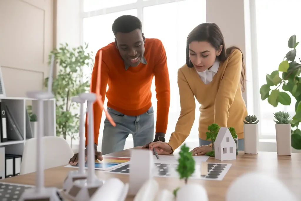 Due giovani, un uomo e una donna, si chinano su un tavolo in un ufficio luminoso, esaminando progetti e modelli legati all'energia sostenibile, inclusi pannelli solari in miniatura e una turbina eolica